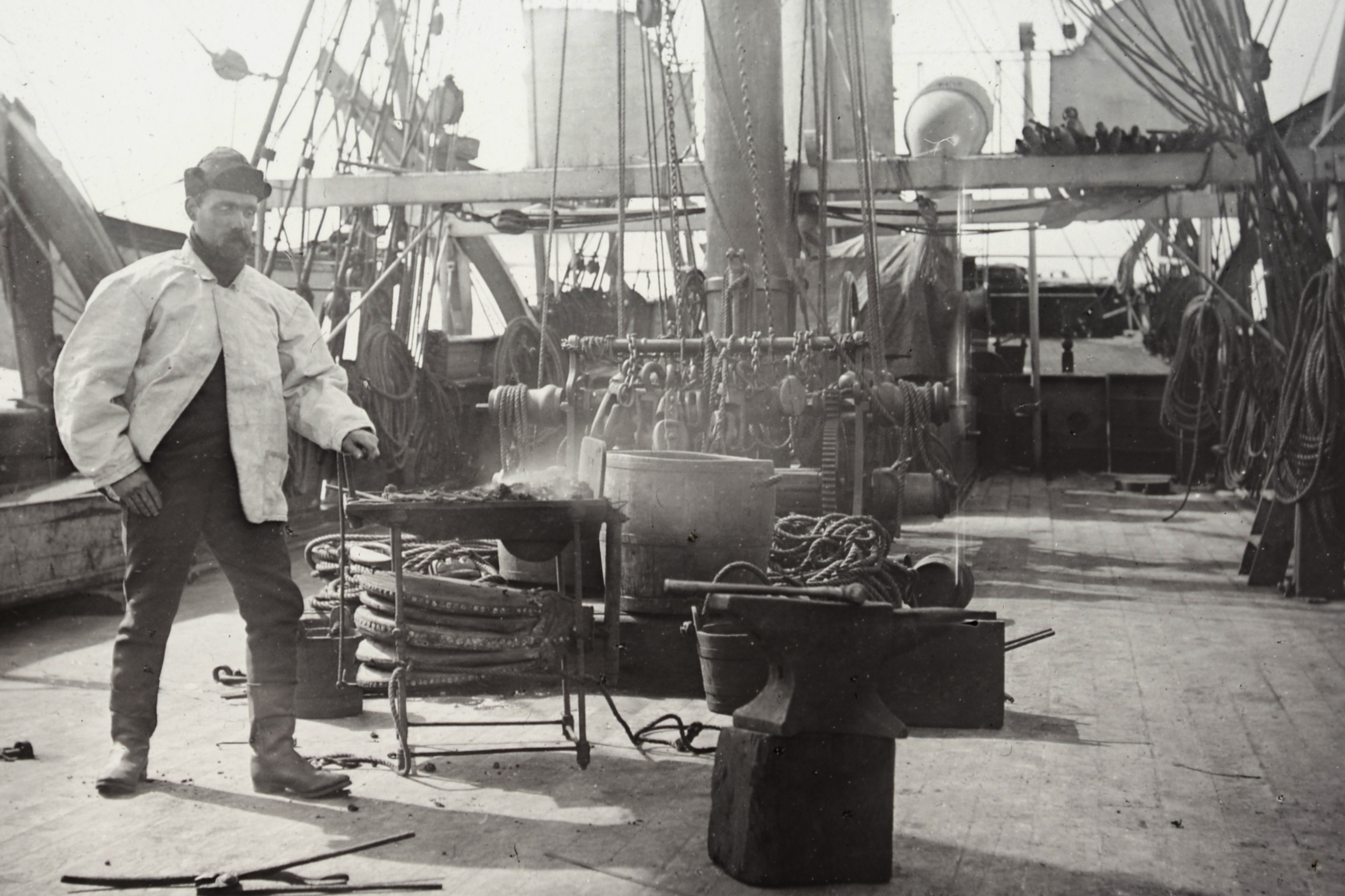 Black and white photograph depicting a blacksmith and his forge on the deck of whaling ship Maud, 1889. In the foreground there are tools scattered on the deck and an anvil with a hammer placed on a block. In the background the deck of the ship is visible with the base of the masts and rigging.