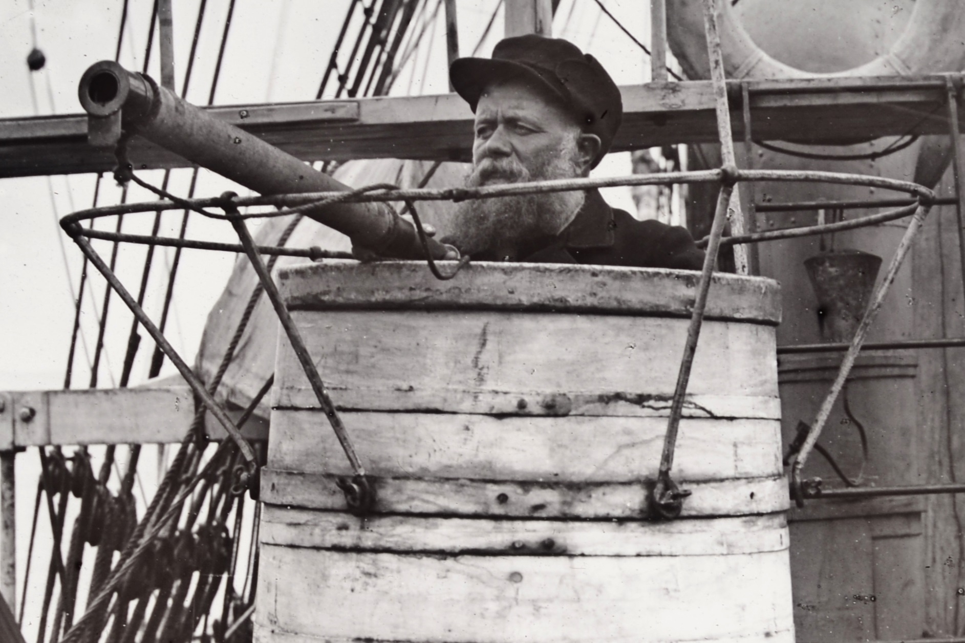 Black and white photograph depicting Captain William Adams, and older man with white beard and a black cap, in the crow’s nest, similar to a white barrel, sitting on the deck of his whaling ship, Maud, 1889. In the background the ship’s deck is visible, with wooden planks, rigging and a ladder.