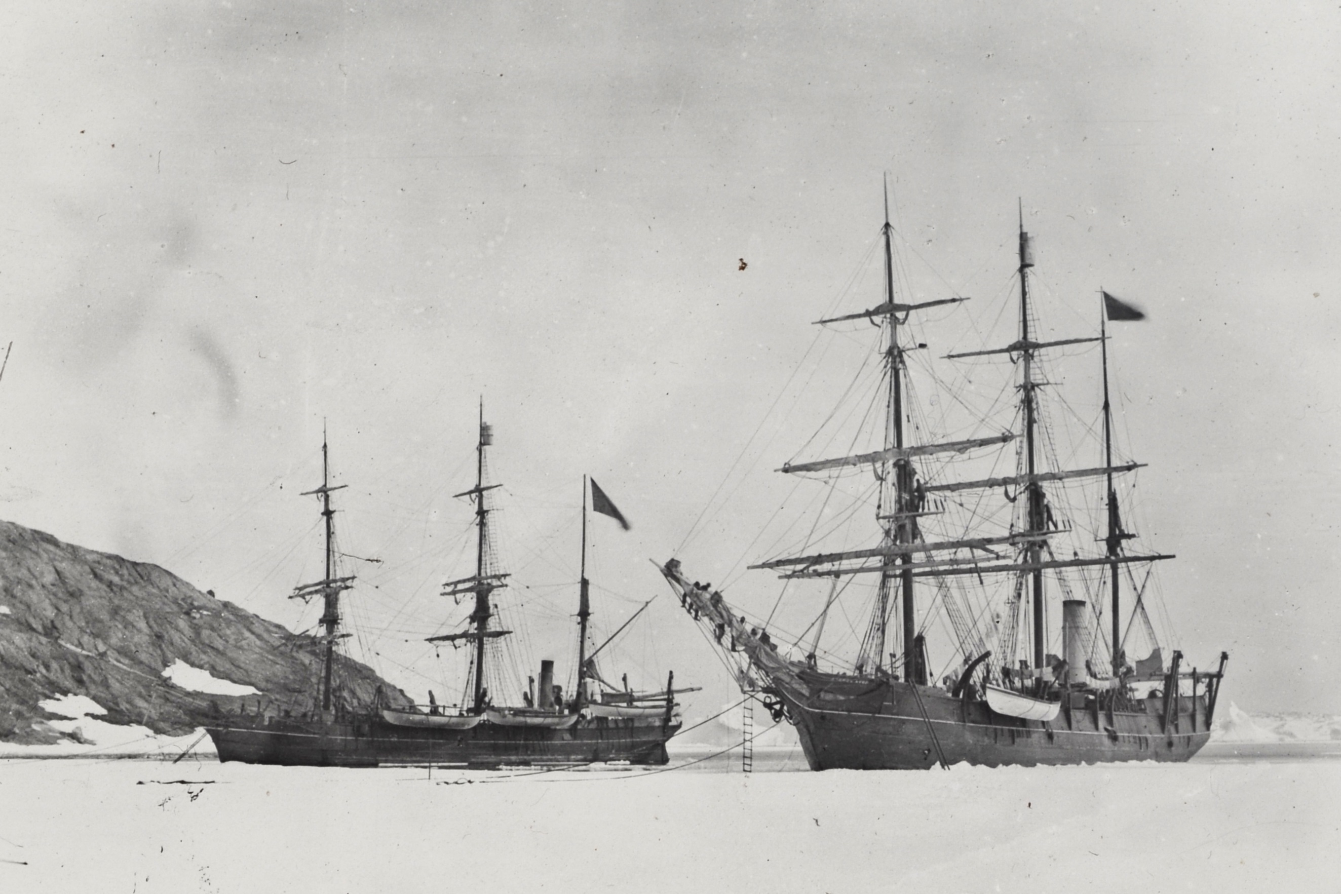 Black and white photograph depicting two sail and steam powered whaling ships, Nova Zembla and Maud, moored to an ice sheet. Men stand in the rigging and on the bowsprit of one of the ships. Rocky coast covered in snow is visible in the background.